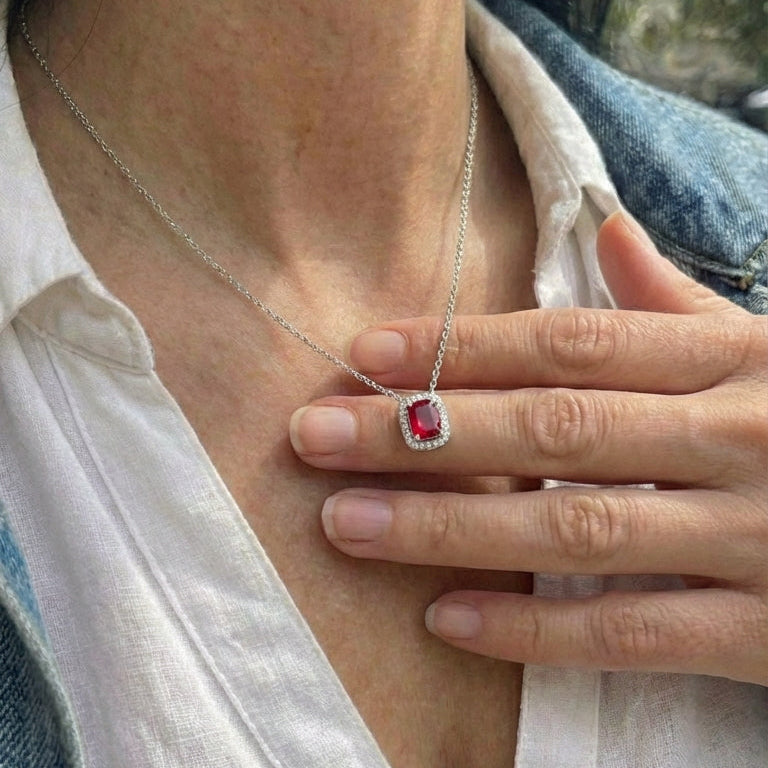 Person wearing a silver necklace with a red gemstone pendant, holding it close to their chest.